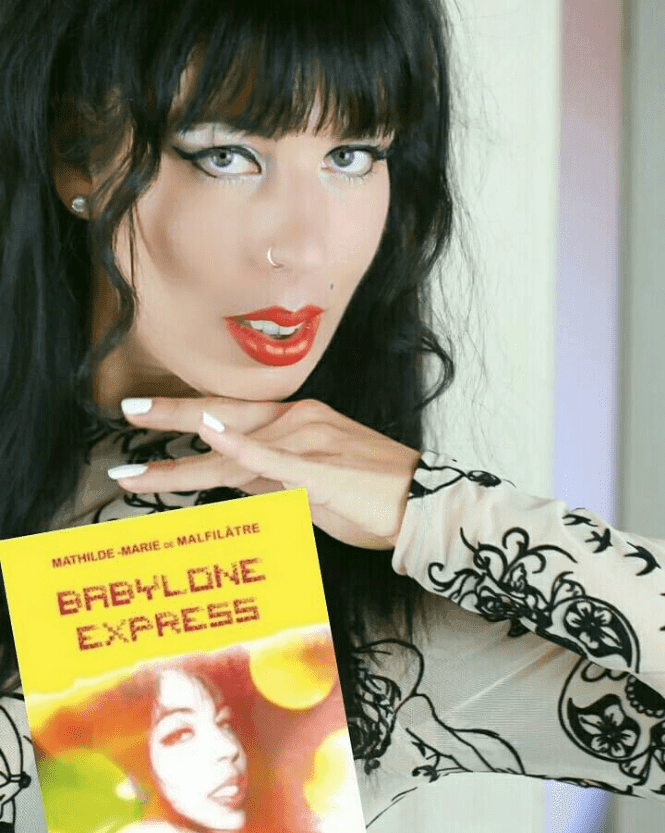 Un portrait en buste de Mathilde-Marie de Malfilâtre avec ses longs cheveux noirs bouclés et son maquillage dramatique. Elle porte un top à manches longues orné d'un motif de fleurs, et tient un livre intitulé "Babylone Express". Son regard est intense et mystérieux.