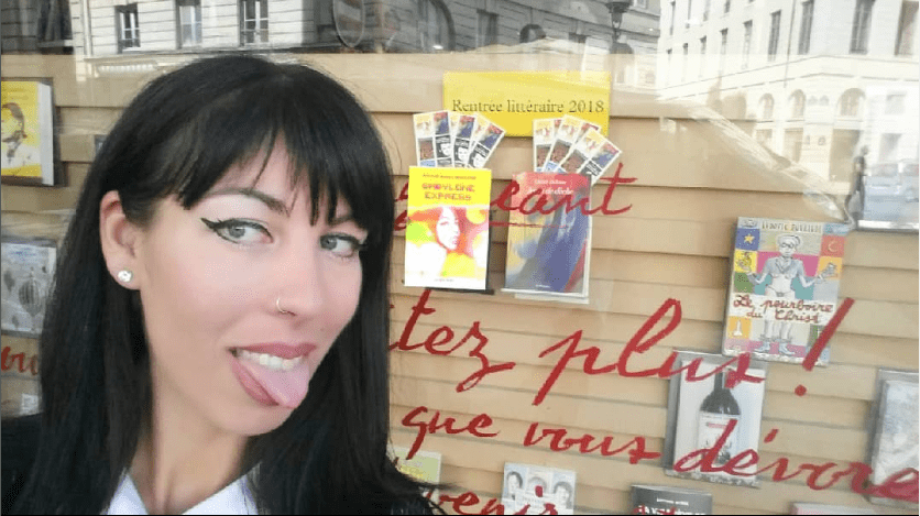 Une photo d'une vitrine de librairie où l'on peut voir plusieurs livres exposés. Derrière Mathilde-Marie de Malfilâtre, qui tire la langue, on distingue un livre intitulé "Babylone Express" avec un portrait de Mathilde-Marie de Malfilâtre sur la couverture.