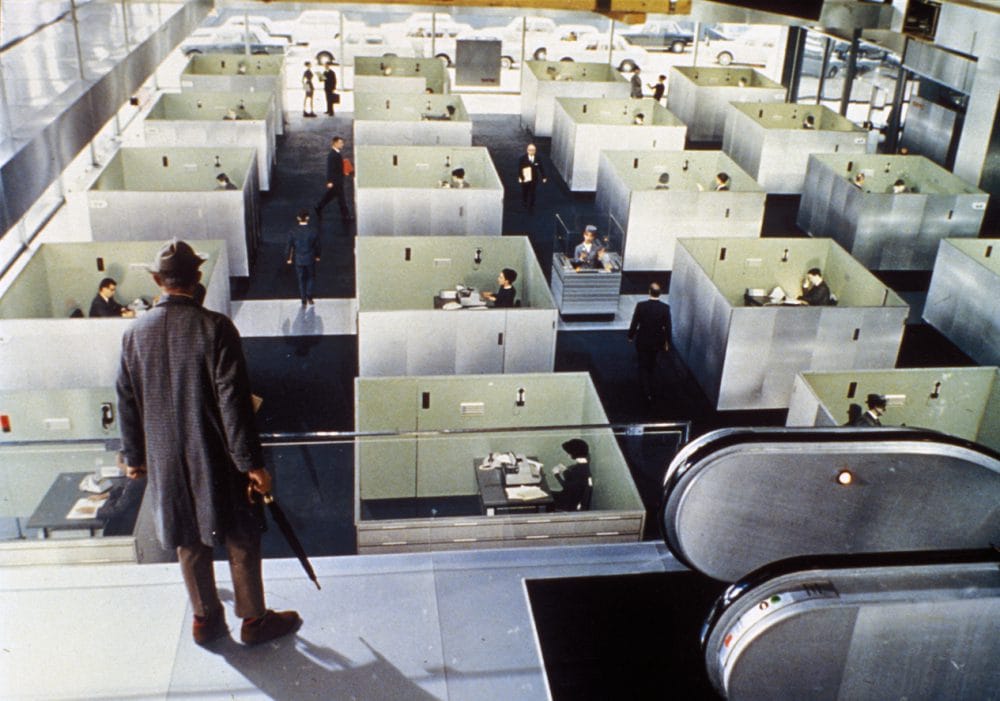 Les Déserts urbains de Jacques Tati, avec un homme de dos avec un chapeau et un parapluie qui regarde, depuis une hauteur, des bureaux carrés séparés les uns des autres.
