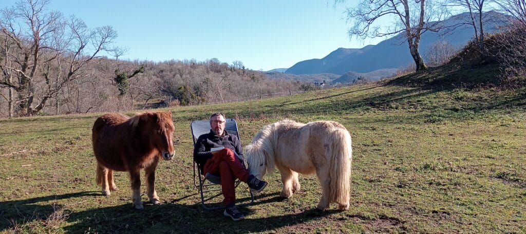 Stéphane Péna entouré de ses poneys et assis sur un sige pliable en pleine nature.